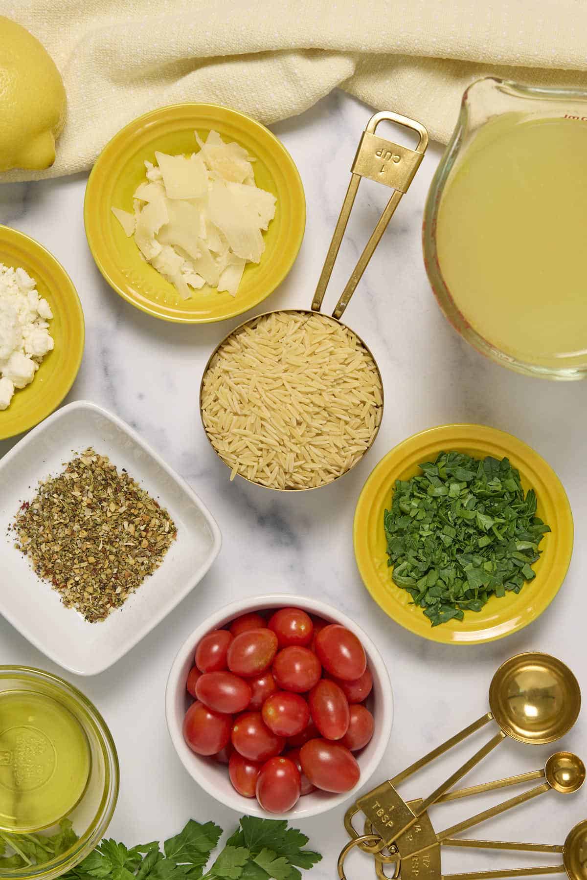 ingredients for the cherry tomato feta orzo recipe; chicken stock, chopped parsley, cherry tomatoes, olive oil, Greek seasoning blend, feta, shaved parmesan, and uncooked orzo
