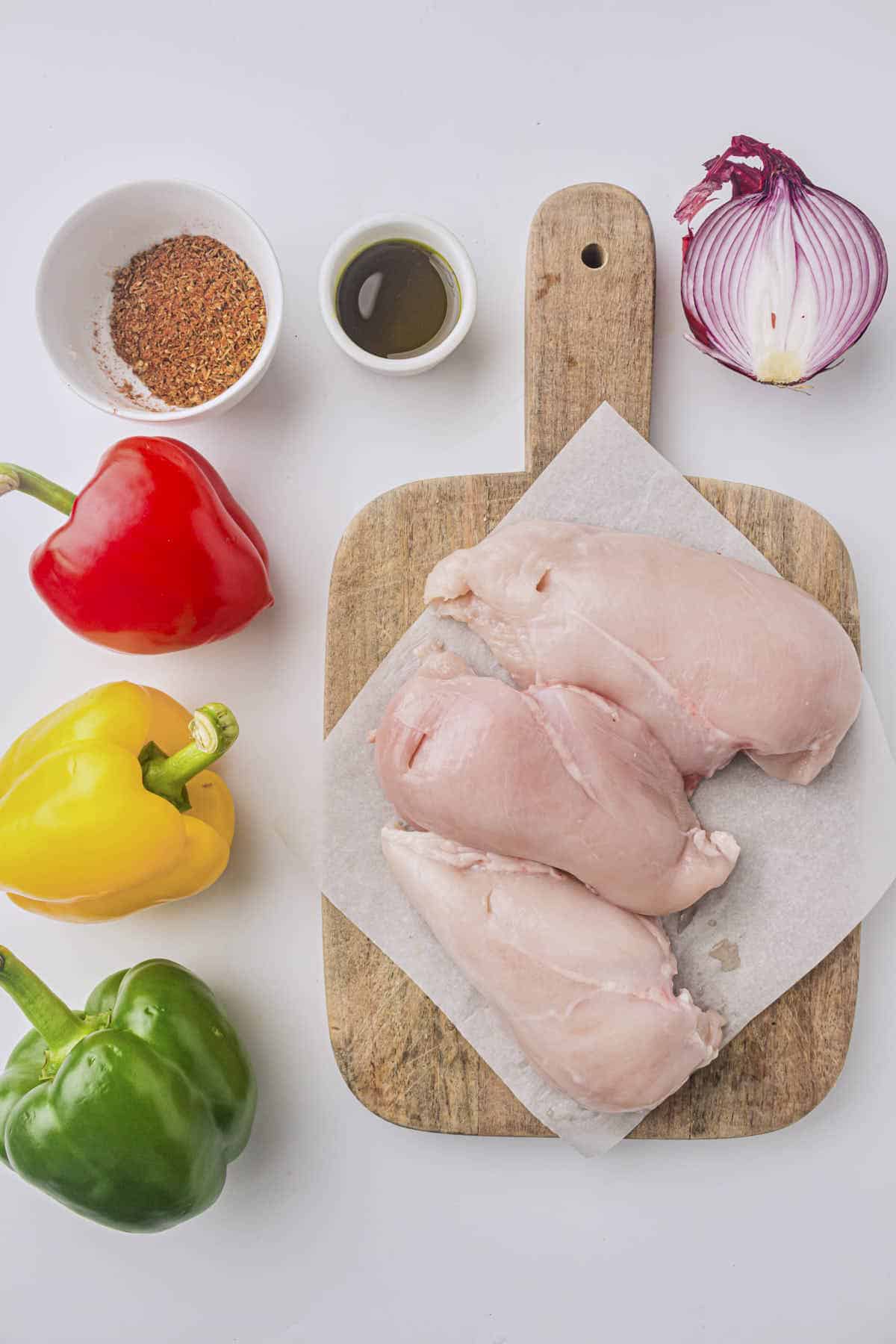 Ingredients for the air fryer chicken fajitas with some seasoning and olive oil in the dishes and chicken on the cutting board with colored bell peppers and half a red onion surrounding