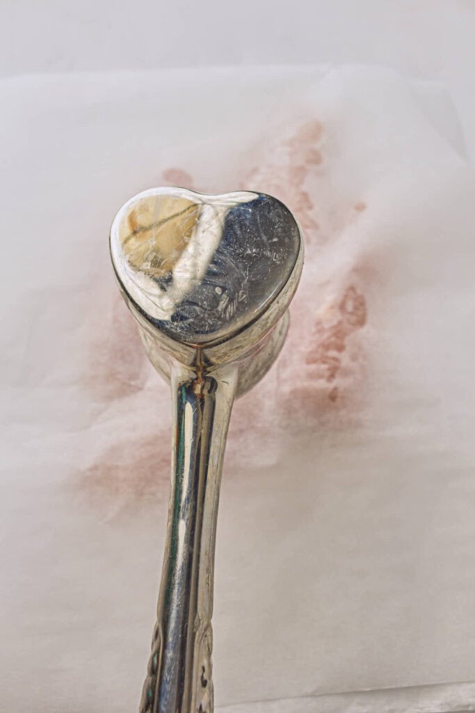 A silver heart shaped tenderizing mallet being used to gently flatten the chicken breasts to make uniformly thick pieces