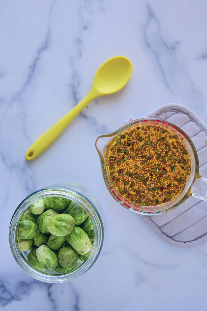 Brussels sprouts in the jar with a measuring cup full of seasoned brine ready to pour in