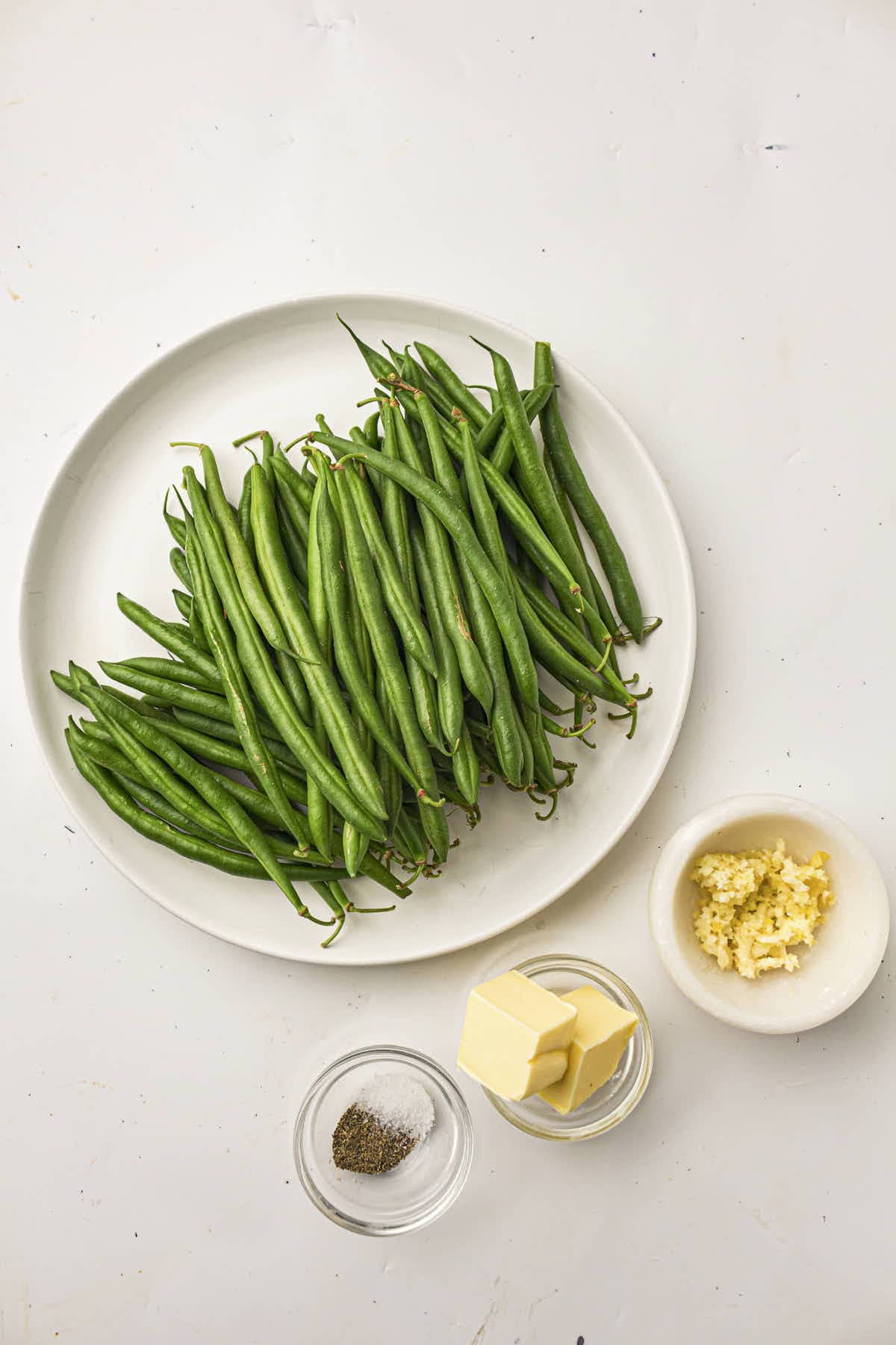 Ingredients for this recipe; a plate of raw green beans with garlic, butter, and salt & pepper in dishes underneath