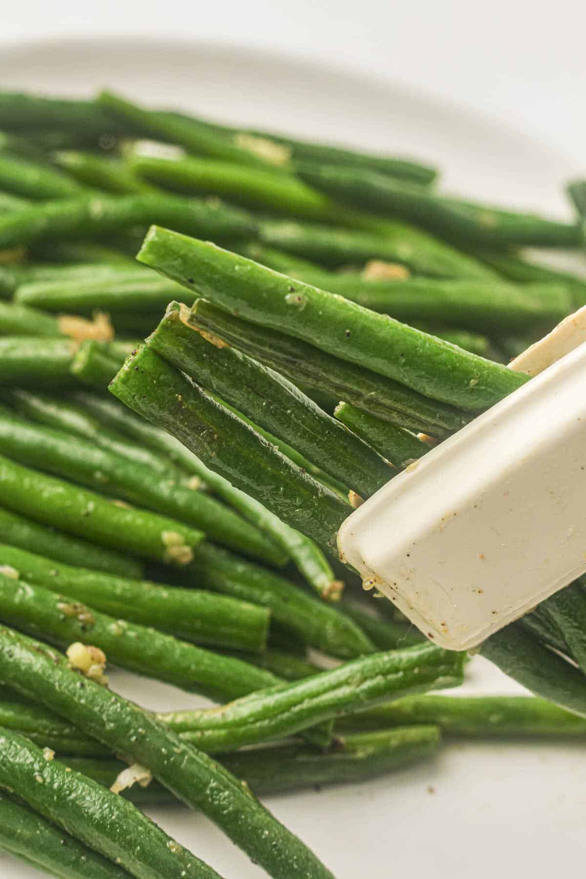 Green beans being held with tongs to show them to the camera with a plate of beans in the background