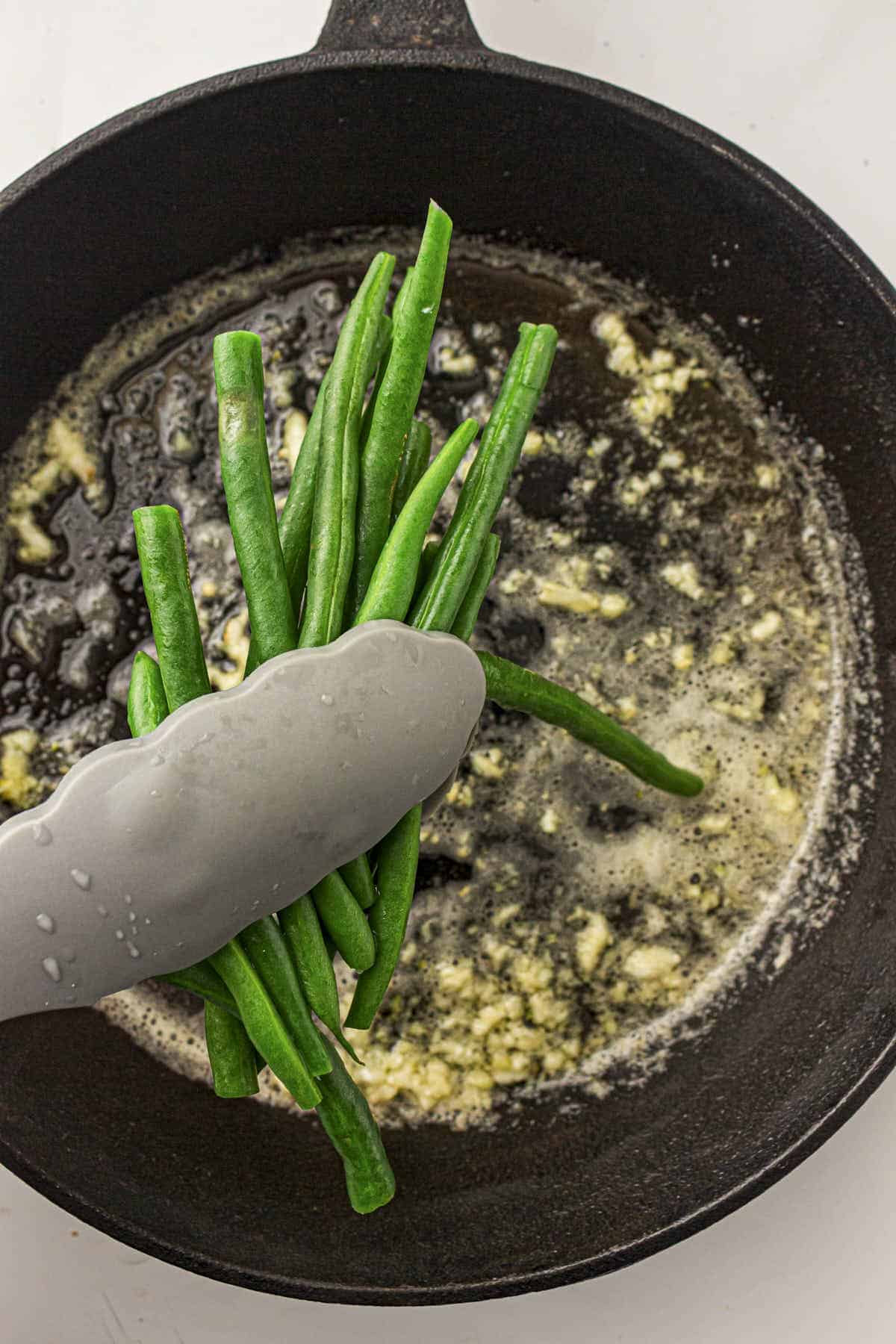 Blanched and dried green beans being placed into the hot cast iron skillet with the melted butter and garlic