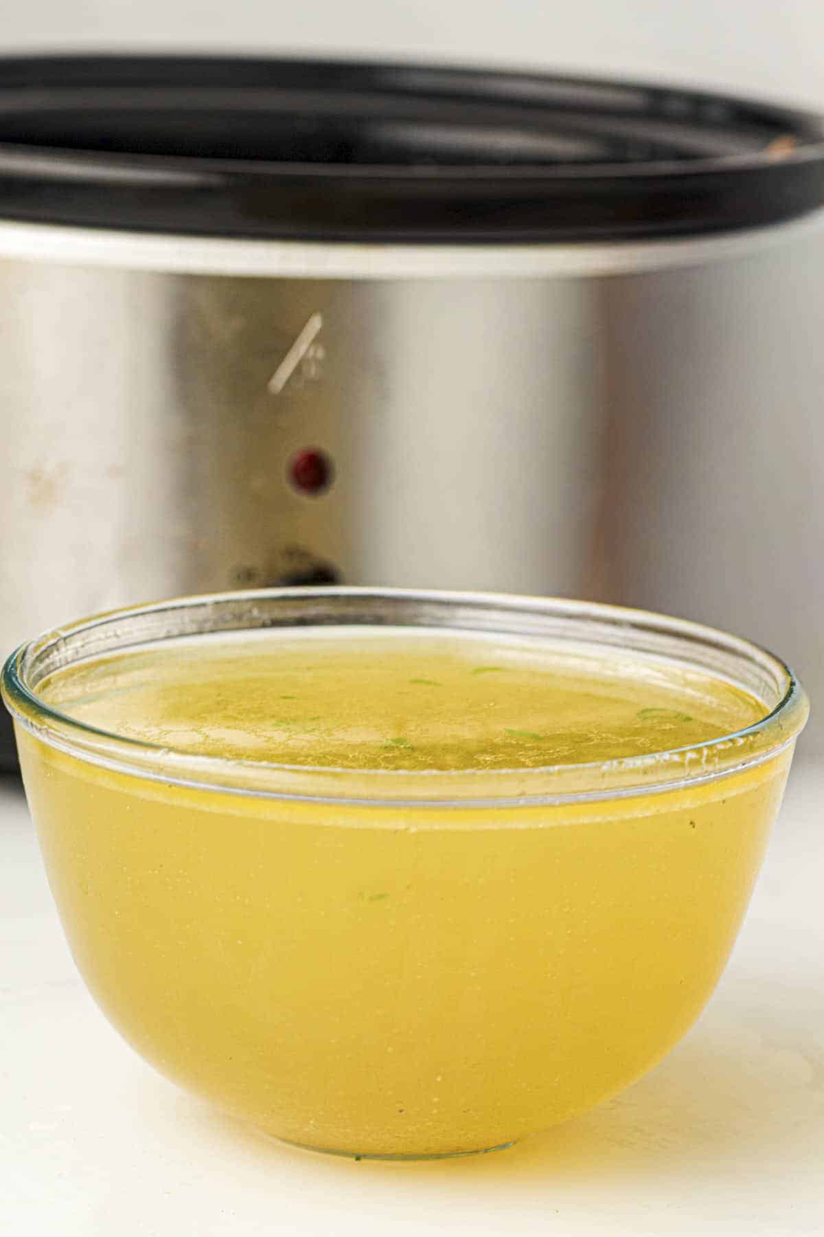 A close up picture of a bowl of chicken stock with a crockpot in the background